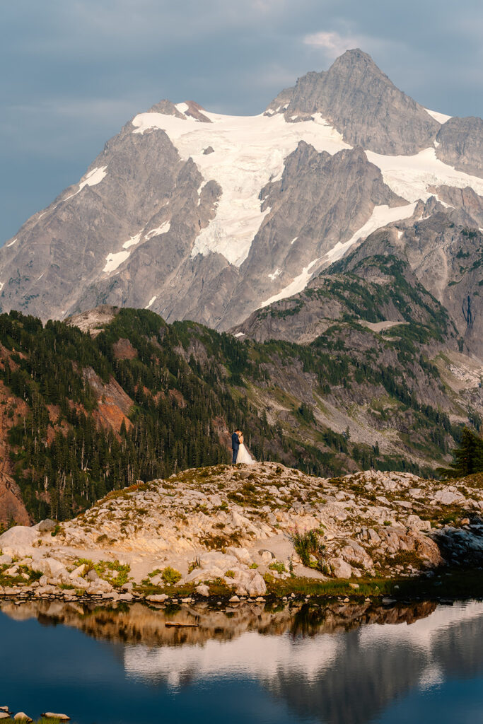 a stunning vertical landscape shot of the couple in their wedding attire. a mountain rises above them and is reflected back in the tarn below them during their mt baker micro wedding 