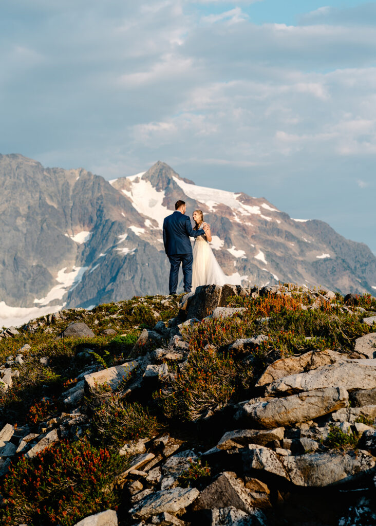 a tender moment of a bride and groom embracing in front of a jagged mountain ridge line with wildflowers blossoming in the foreground 