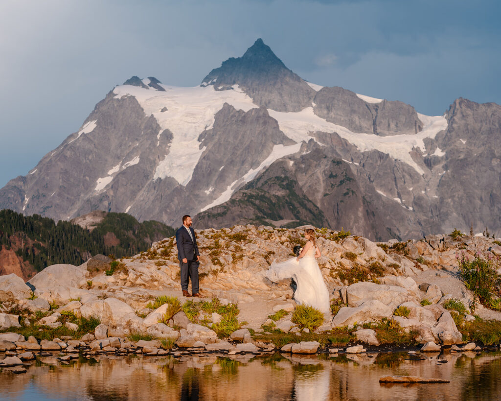 a bride swishes her wedding dress for her groom. mountains rise behind them as they gaze at each other during their mt baker micro wedding 