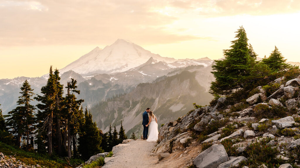 a large landscape shoing mt baker in the throws of golden hour with a bride and groom following a trail towards it during their mt baker micro wedding 
