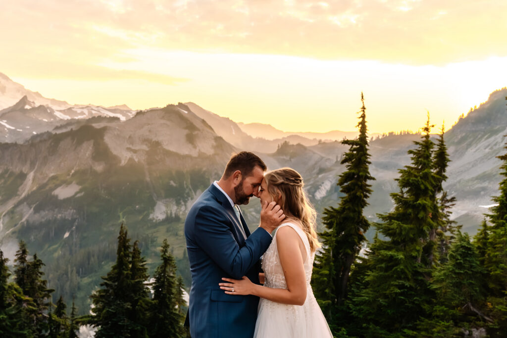 a bride and groom press their foreheads together in a serene moment. beyond them is a stunning mountain range highlighted by a glowing sunset 