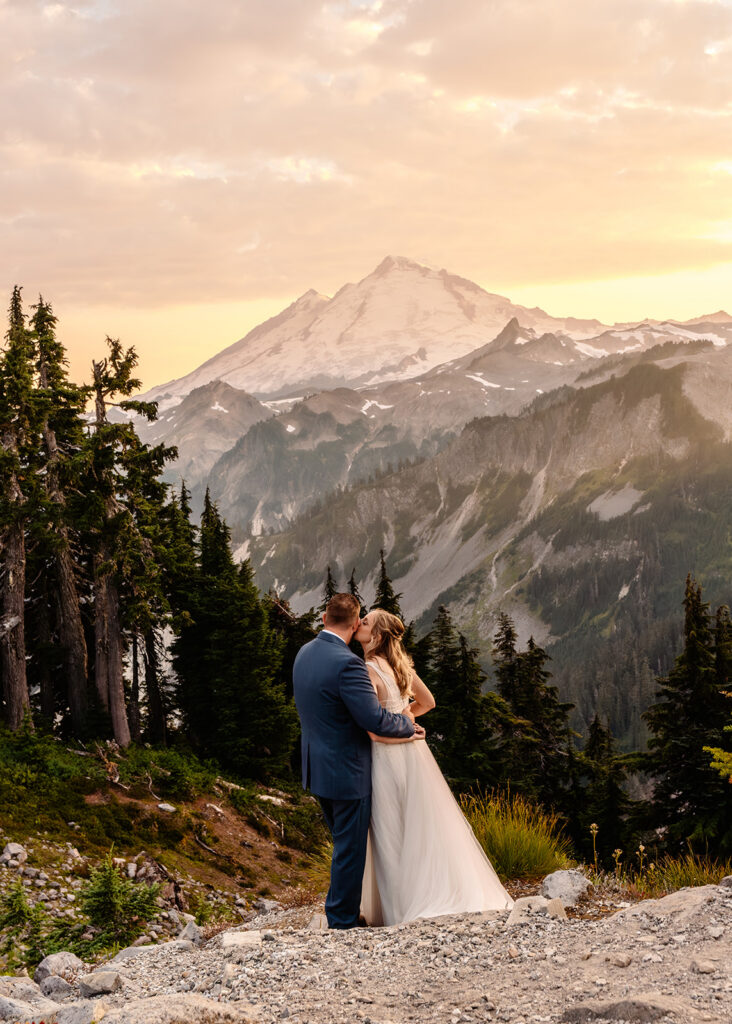 a bride and groom in their wedding attire gaze out at a stunning mt baker during a golden pink sunset. The shot is from begind them. The bride kisses her grooms cheek during their mt baker micro wedding 
