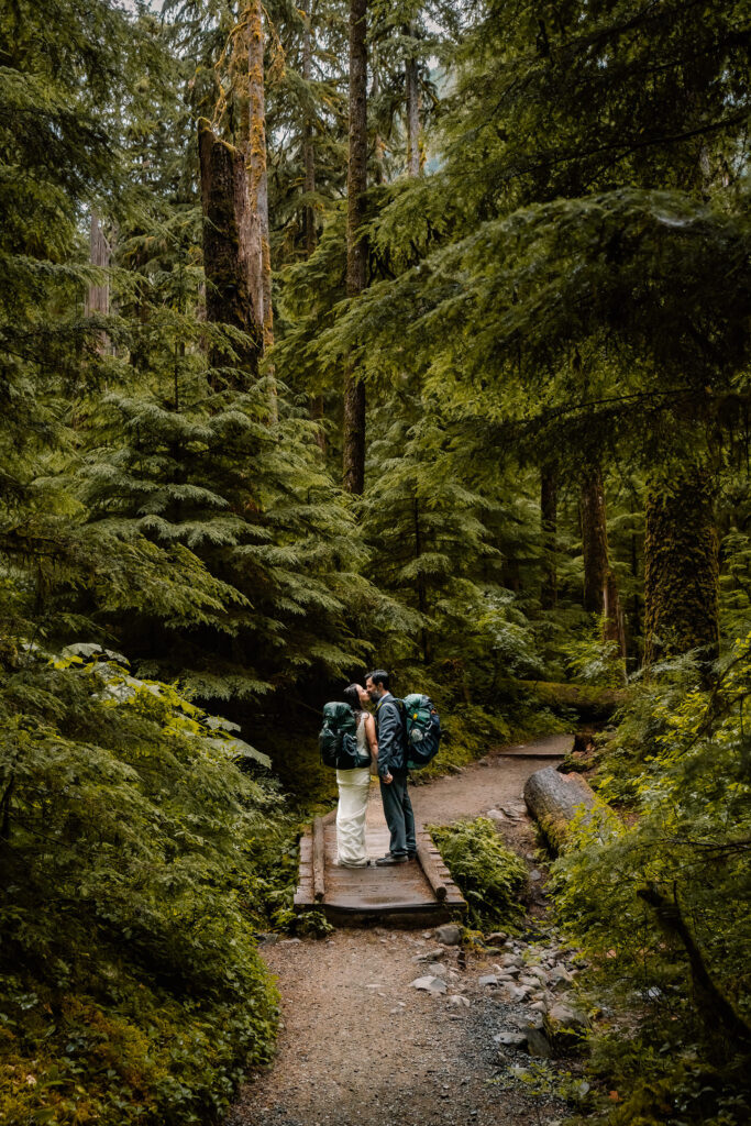 a couple in their wedding attire and hiking backpacks kiss a on a small wooden bridge as they explore the old growth forest for their elopement 