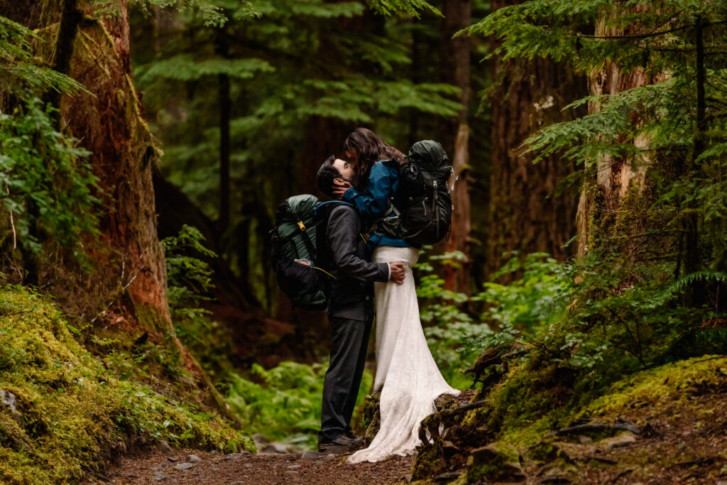 a couple in their wedding attire, with hiking backpacks secured kiss in the middle of a forested trail during their rainforest elopement 