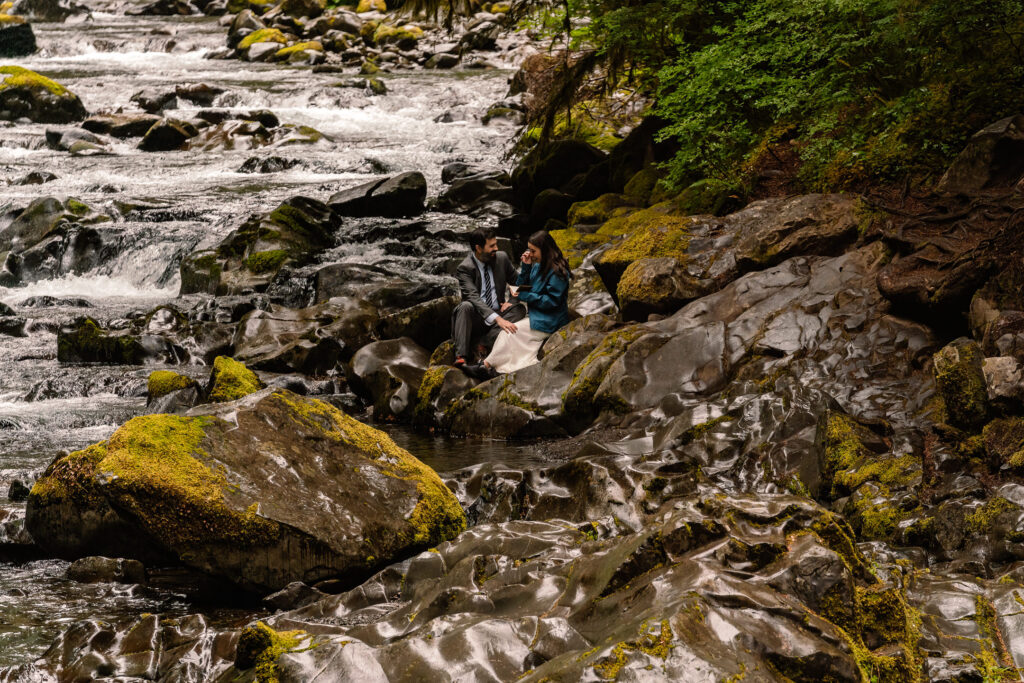 a couple in their wedding attire, sit on the rocks of a rushing sol duc river as they exchange vows 
