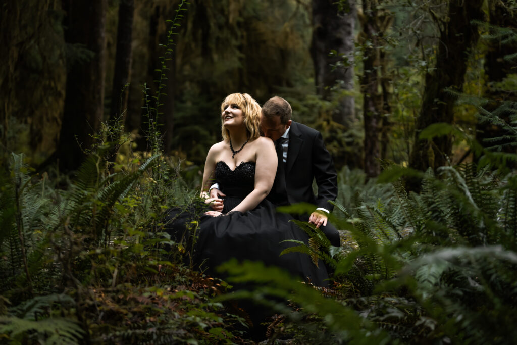 a couple in all black wedding attire sit in the middle of a group of ferns. The scene is moody and lush during their hoh rainforest elopement