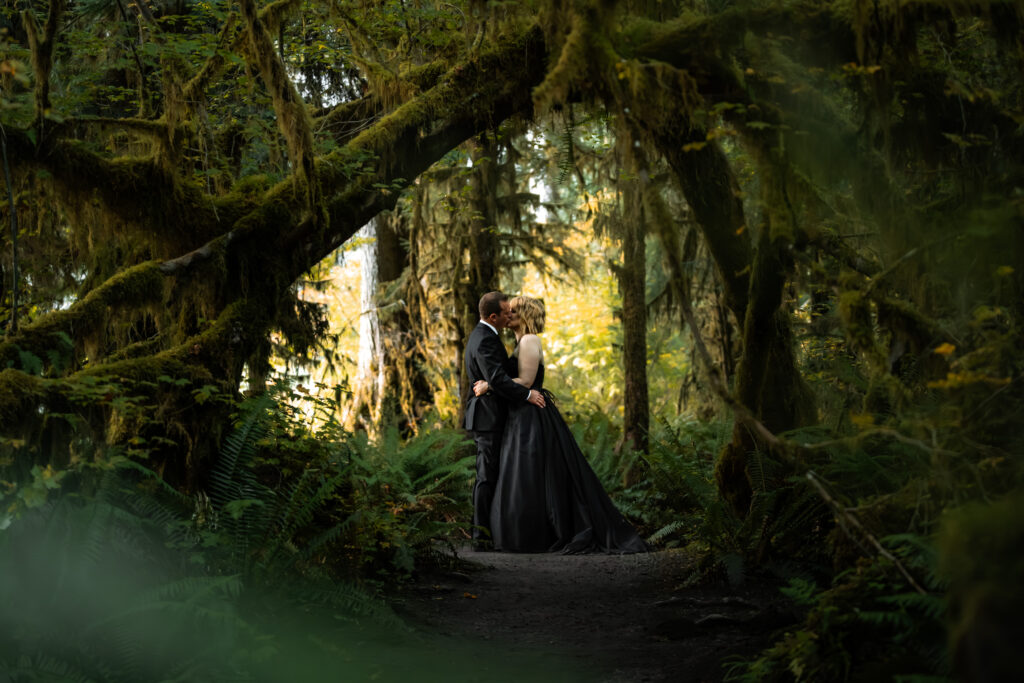a couple in all black wedding attire embrace under a thick, mossy canopy during their hoh rainforest elopement