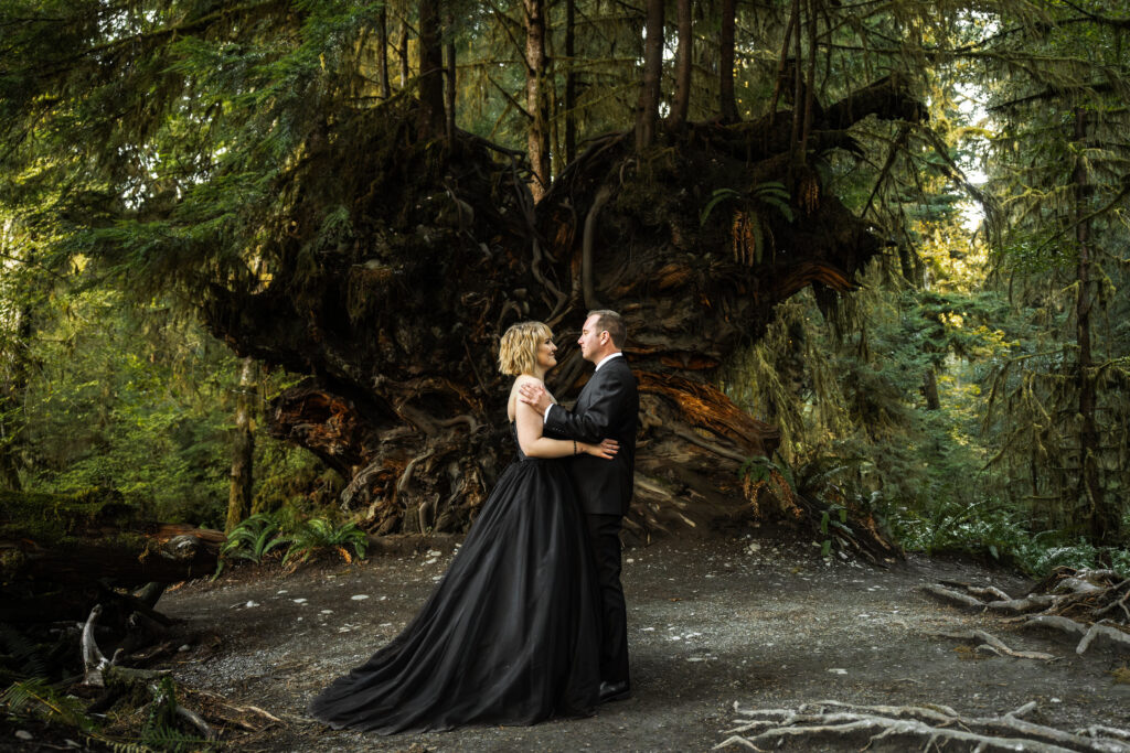 a couple in all black wedding attire embrace in front of a massive root system that is shaped like a heart during their Hoh Rainforest elopement