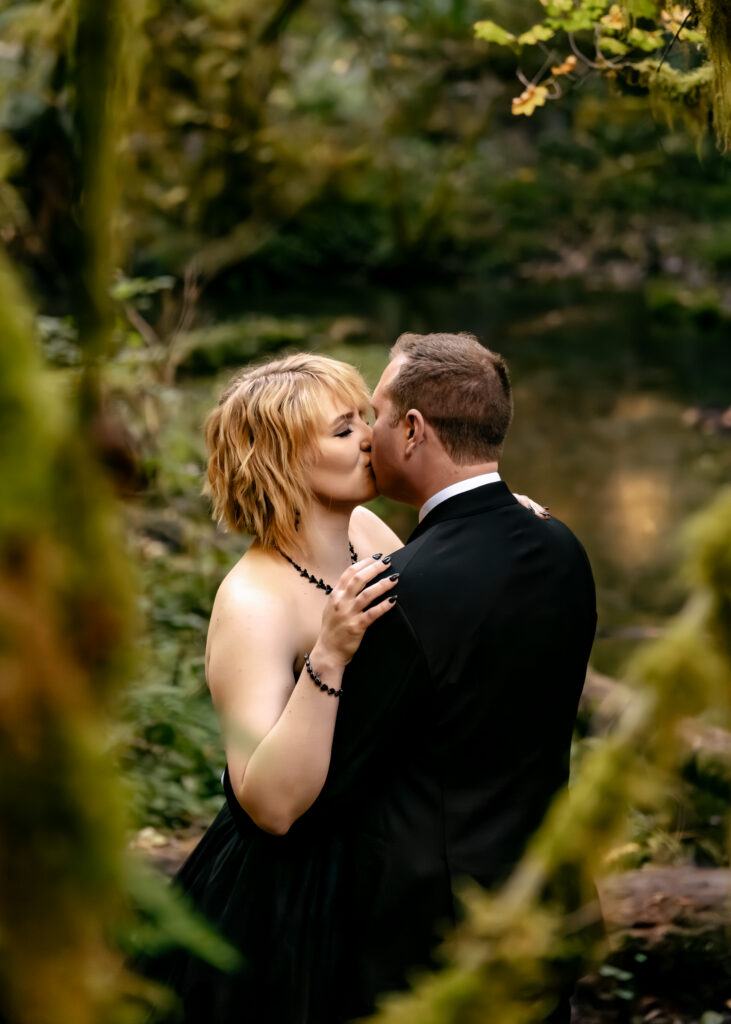 a couple in all black wedding attire kiss. They are framed by green moss during their hoh rainforest elopement