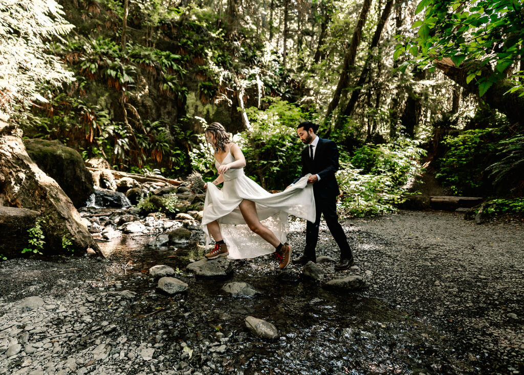 a couple in their wedding attire cross a small creek. Our bride leads the way as her groom carries the train of her wedding dress 
