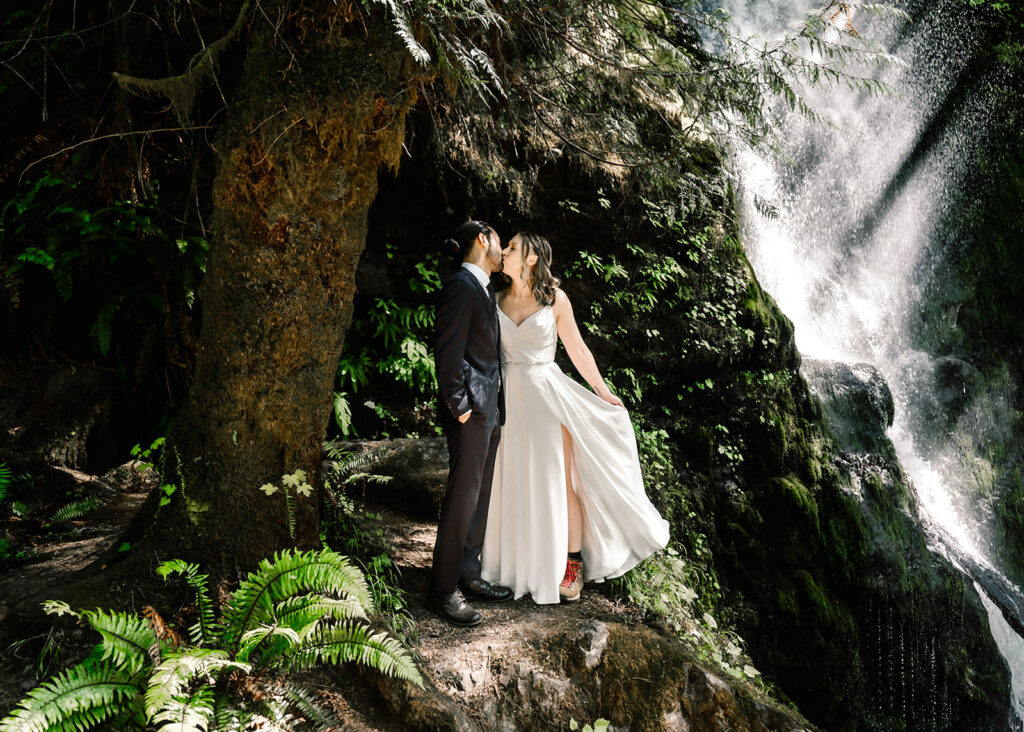 a couple in wedding attire share a casual kiss in the middle of a verdant forest beside a shimmering waterfall 