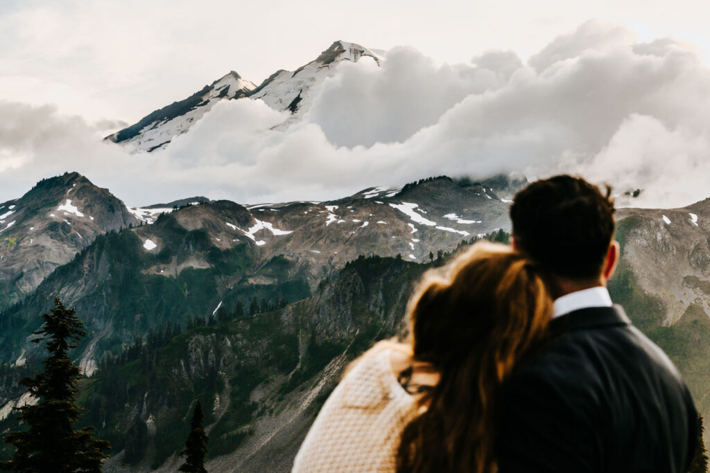 a bride and groom paint the foreground. They are blurred as the bride rests her head on her grooms shoulder. We see their perspective. they gaze out at a stunning Mt. baker painted with clouds 
