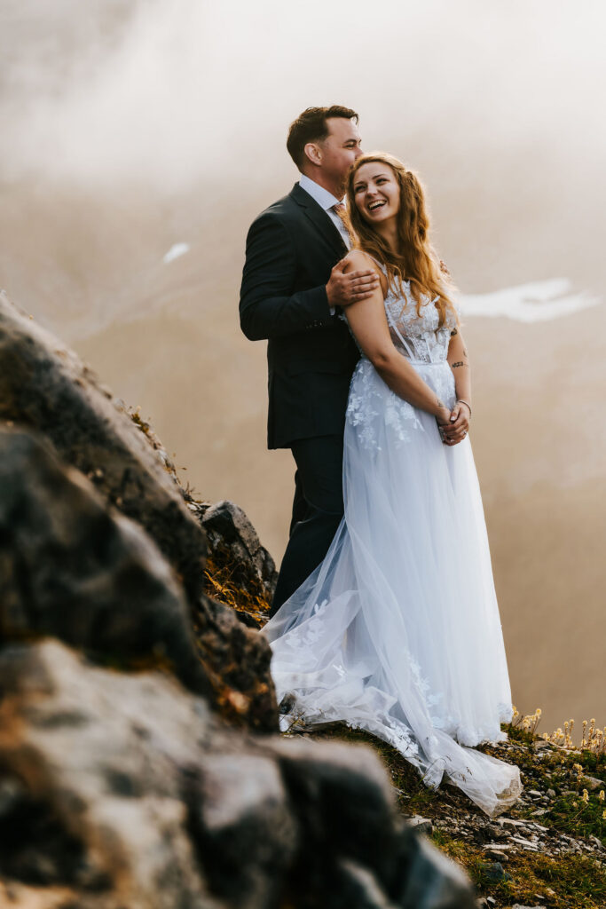 a groom holds his bride as she peers back at him. The landscape before them is a mix of gold and green smeared with pearly white clouds