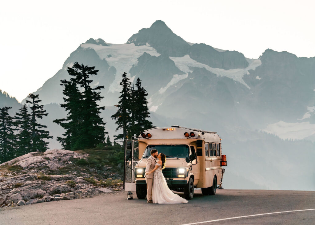 a couple in their wedding attire embrace in front of a bus that they live in, full-time. A stunning mountain scene fills up the background for their artist point elopement 