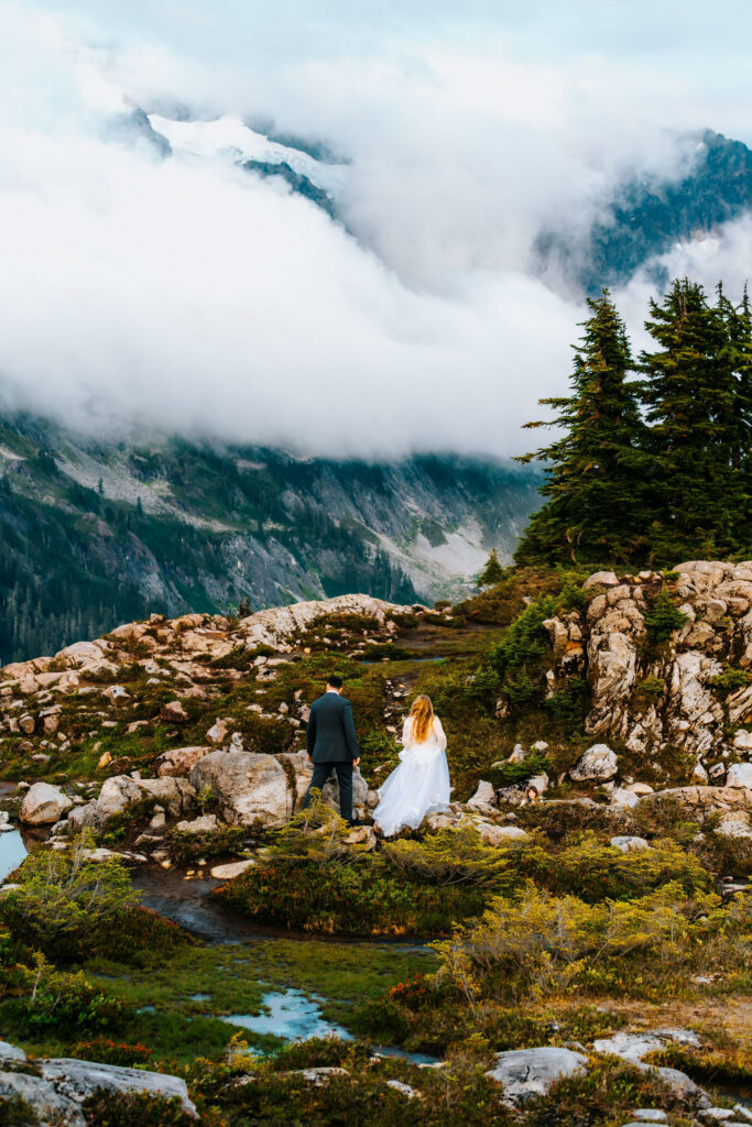 a bride and groom explore the landscape that is dominated by deep greens and blues. Mountains are smeared with clouds as they search for the perfect spot for their artist point elopement