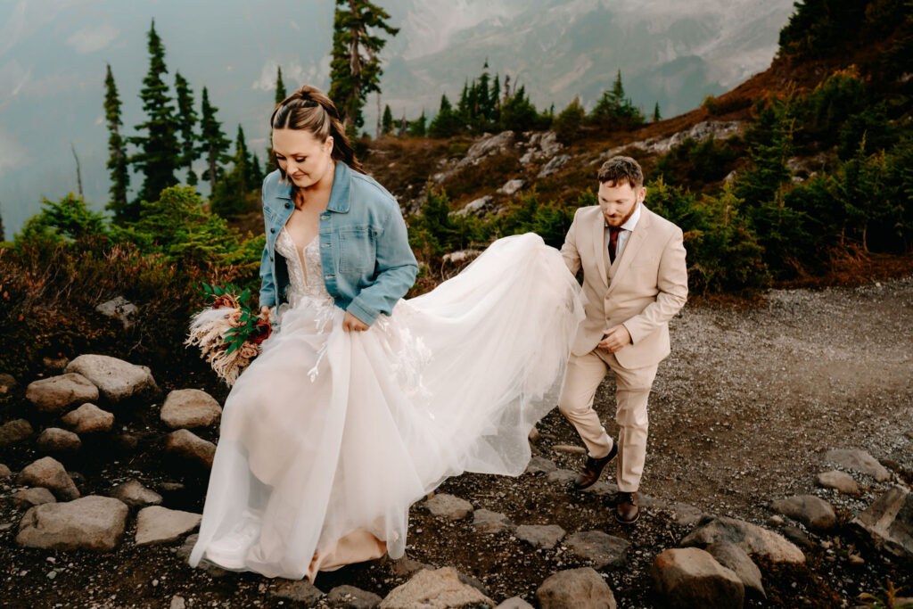 a bride and groom hike to find the perfect location for their artist point elopement. As they hike, the bride leads as her groom carries the train of her wedding dress
