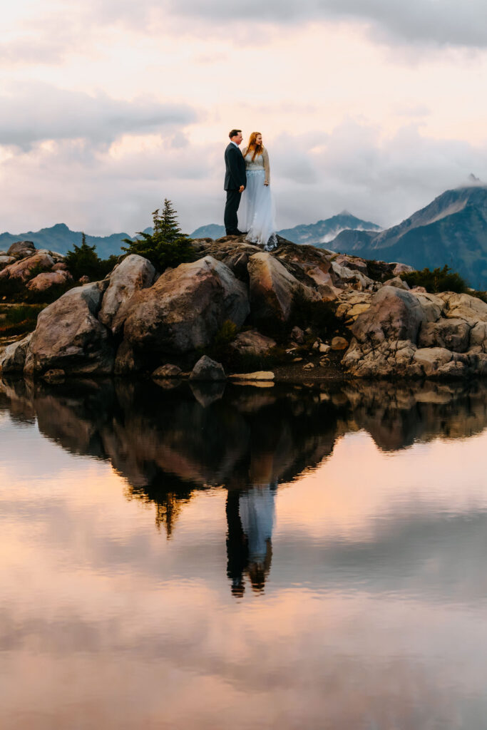 a bride and groom stanf of a rocky surface. The brilliant sunset is reflected in the tarn below them 