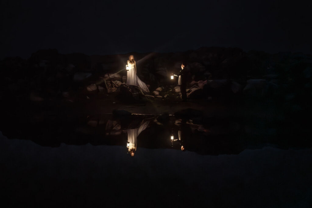 a moody, night time shot of a bride and groom. They hold lanterns and are perfectly reflected in the still waters of the tarn below them following their artist point elopement