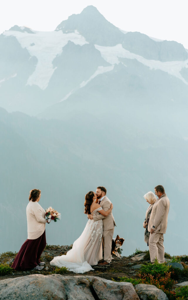a couple in their wedding attire are surrounded by family members as they share a first kiss during their artist point elopement. Blue, snowy mountains rise behind them and soft, morning light paints the scene