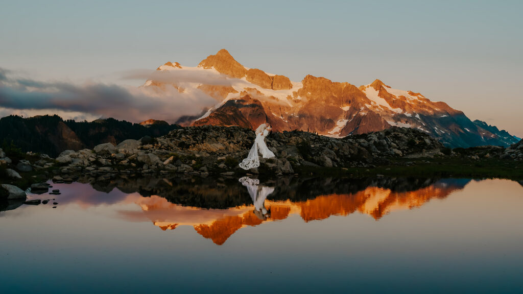 a stunning landscape shot of orange pink mountains lit by alpine glow. a couple in their wedding attire dip kisses on a rocky surface as they're reflected in the still tarn below them during their artist point elopement