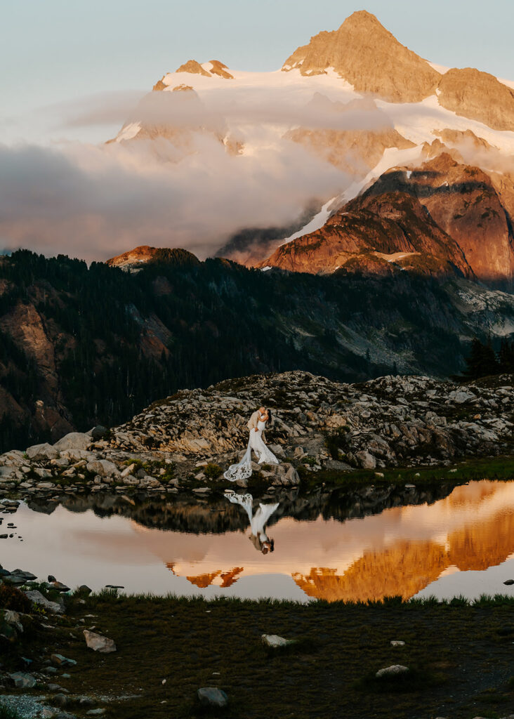 a couple dip kiss in front of an impressive mountain peak in their wedding attire. a reflective tarn mirrors below them during their artist point elopement 