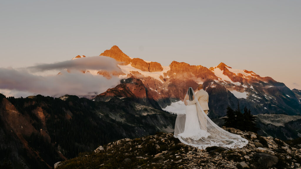 a couple in their wedding attire gaze out at the mountains before them. they glow with the setting sun. The brides veil spreads behind her as they embrace during their artist point elopement 