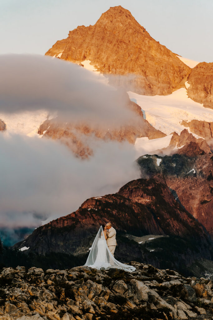 a couple in wedding attire kiss in front of a staggering mountains glowing with the setting sun during an artist point elopement