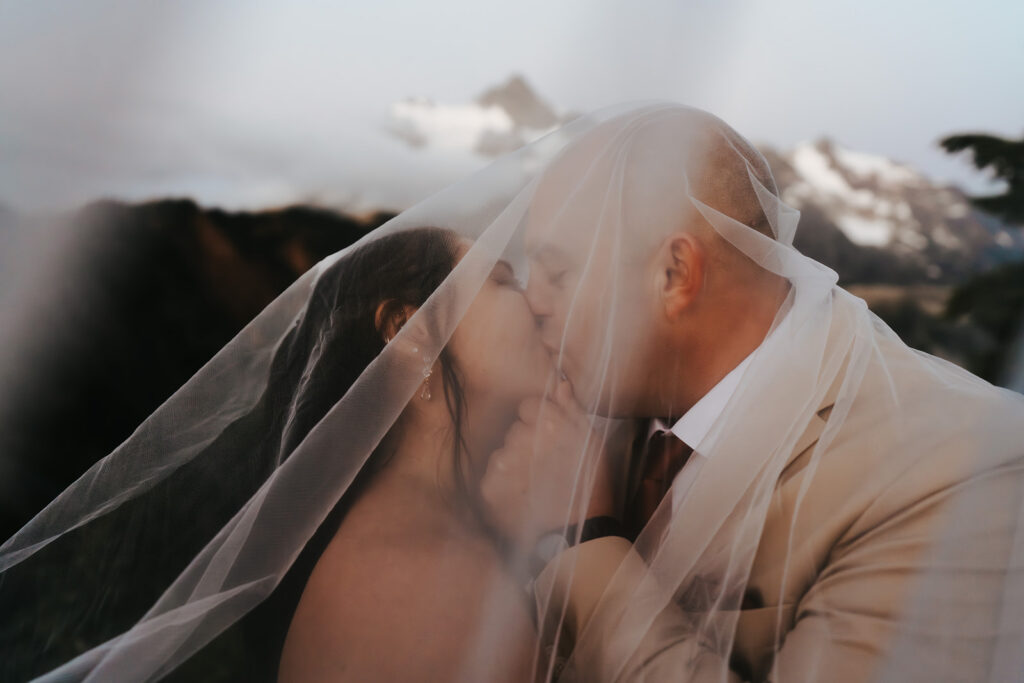 a groom deeply kisses his bride under her veil. mountain peaks stand out behind them during their artist point elopement