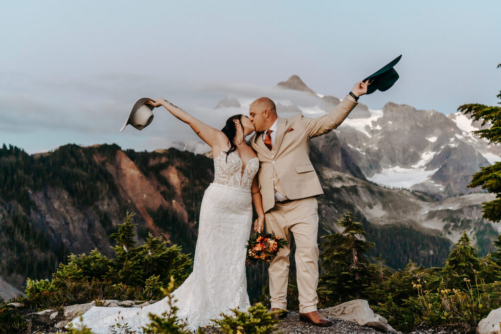 a bride and groom kiss and toss their cowboy hats during their artist point elopement. mountains frame their background as they celebrate 