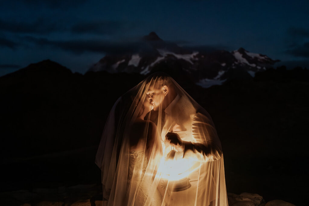 a couple in wedding attire kiss deeply under the brides veil. they are lit only by the glow of their lantern. a snowy mountain can be seen faintly behind them during their artist point elopement 