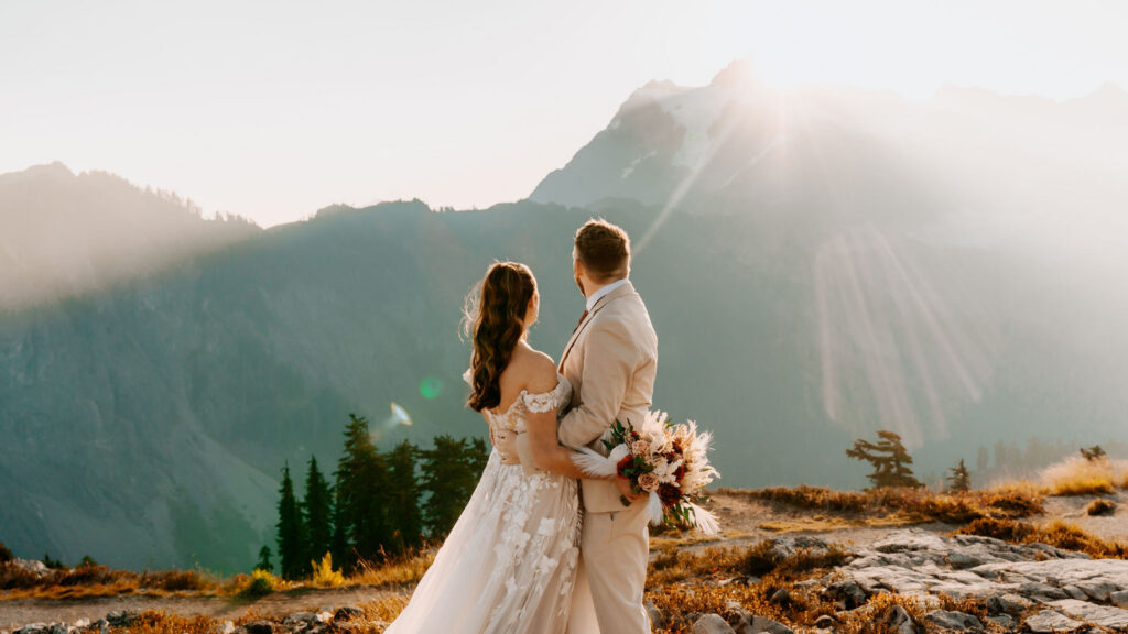 a couple gazes out at the mountain landscape as the sun rises from behind the mountains. Light leaks through the frame giving a magical feel to this artist point elopement 