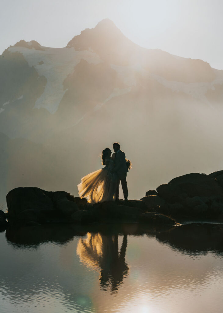 a bride and groom are silhouetted in front of a mountain ridge line sunlight rises and the tarn below them sparkles with their reflection and golden light 