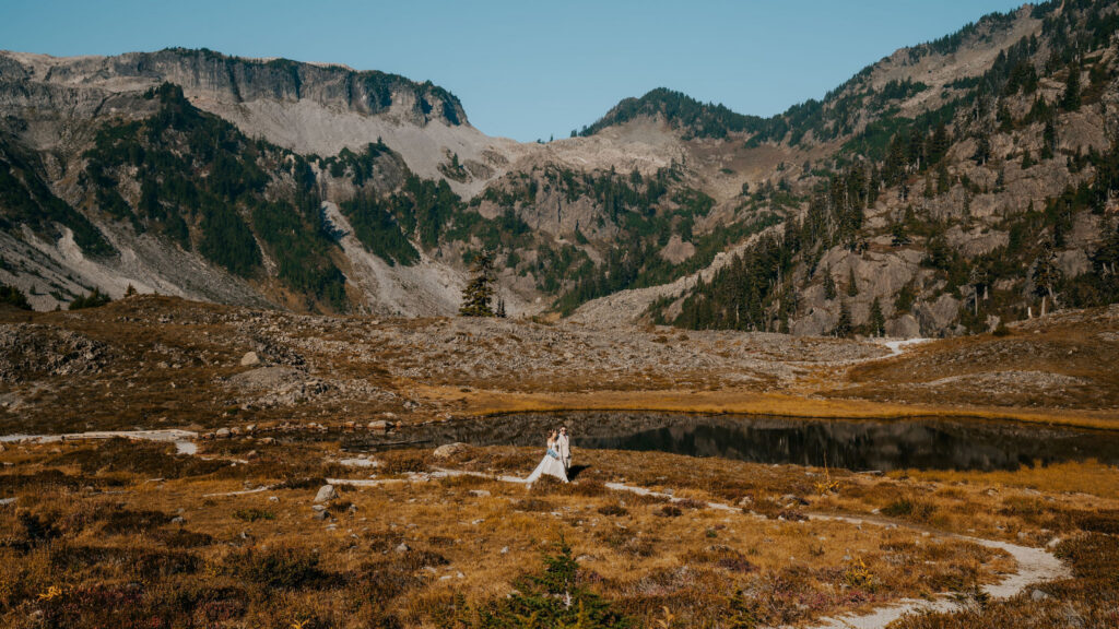 a couple, in their wedding attire explore a mountain terrain after their artist point elopement. The landscape is turning to autumn colors, and the sunlight is high above them 
