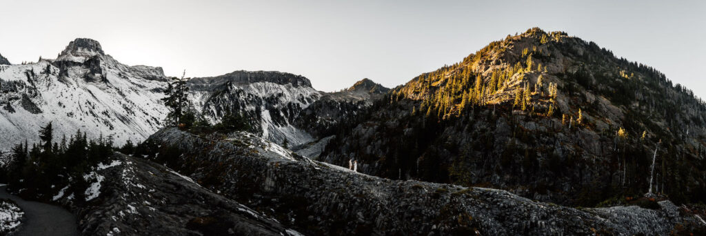 a panoramic shot of a rocky, mountain landscape. A bride and groom are perfectly centered between shadow and light. They are teeny tiny in the massive landscape 