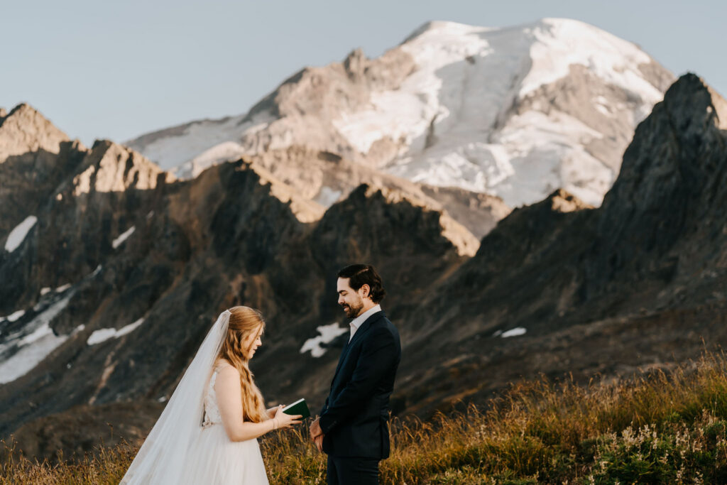 a bride and groom stand in their wedding attire in front of a massive Mt. Baker. The bride, wearing a long flowing veil tearfully reads her vows to her groom, who beams with joy.