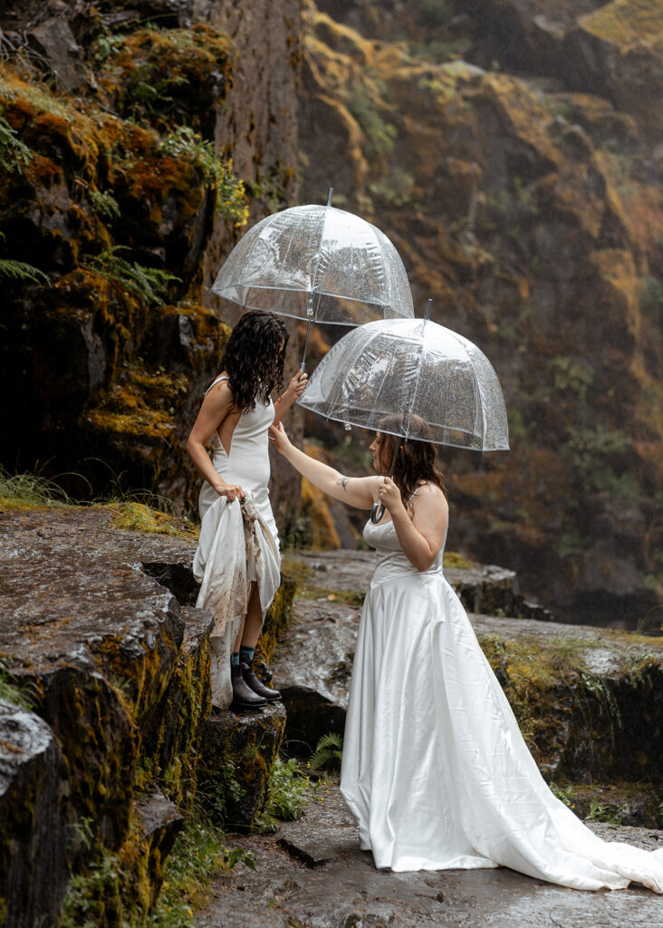 two brides hike down a rocky cliff with clear umbrellas in their wedding dress 