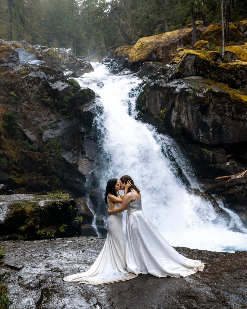 two brides kiss in front of a gushing waterfall