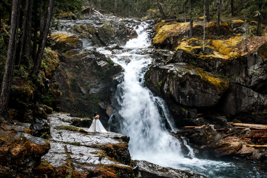 two brides embrace in a wide landscape shot of a gushing waterfall. the basalt rocks are covered in a bright green moss 