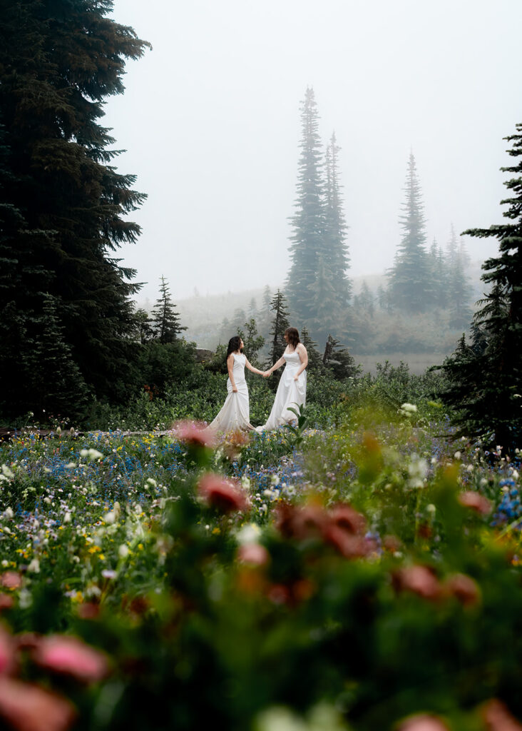 two brides walk through a meadow of wildflowers in a foggy landscape during their tipsoo lake elopement 