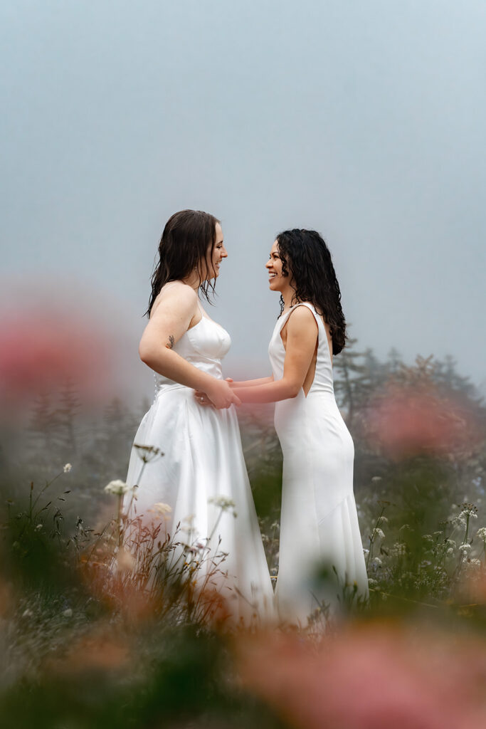 two brides embrace and smile broadly at each other. they are framed by pink wildflowers 