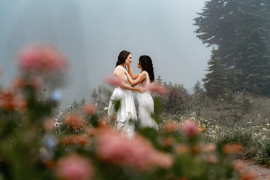 a bride touches the cheek of her bride. they are framed by pink wildflowers 