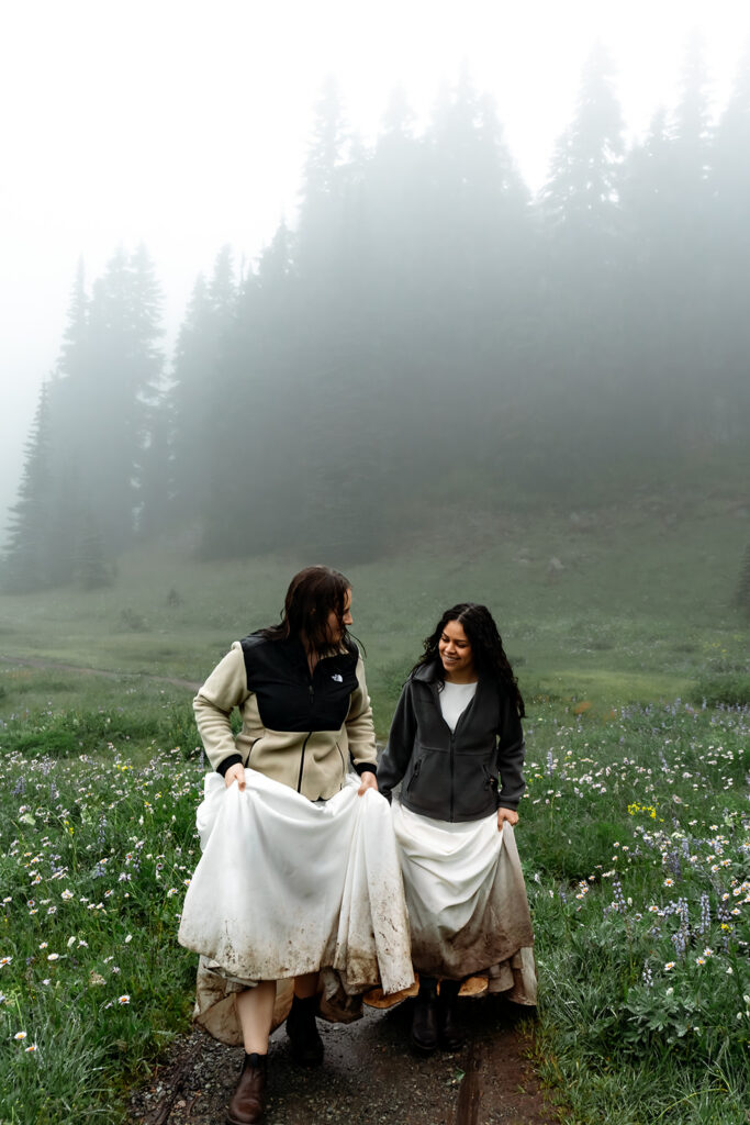 two brides explore the wildflower meadows of tipsoo lake during their elopement. the bottoms of their dresses are muddy and they smile broadly at each other as they hike 