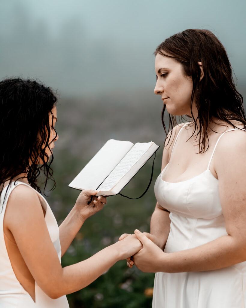 a bride reads a book of poems to her bride as they hold hands and gaze lovingly at each other 