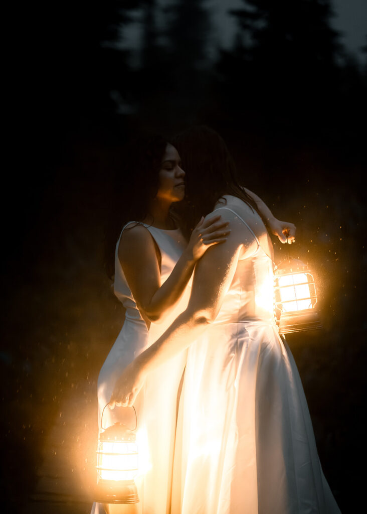 two brides embrace and are lit only by warm lantern light rain falls around them and they smile sweetly 