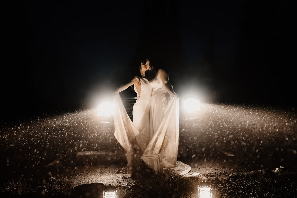two brides kiss in front of car head lights as they dance. the rain looks like sparkles in the darkness of the night 