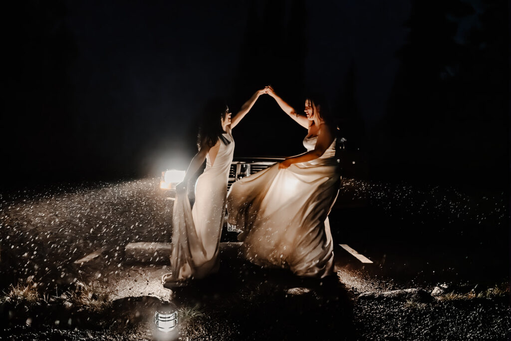 two brides kiss in front of car head lights as they dance. the rain looks like sparkles in the darkness of the night 
