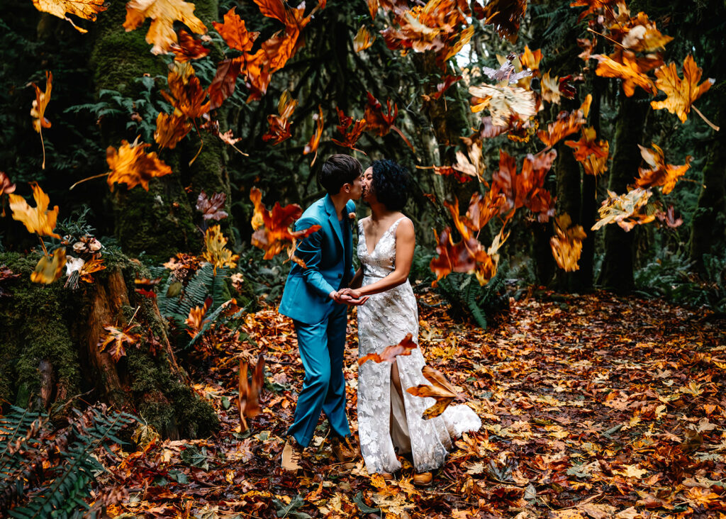 a couple in their wedding attire kiss as bright autumn leaves fall around them in the shape of a heart