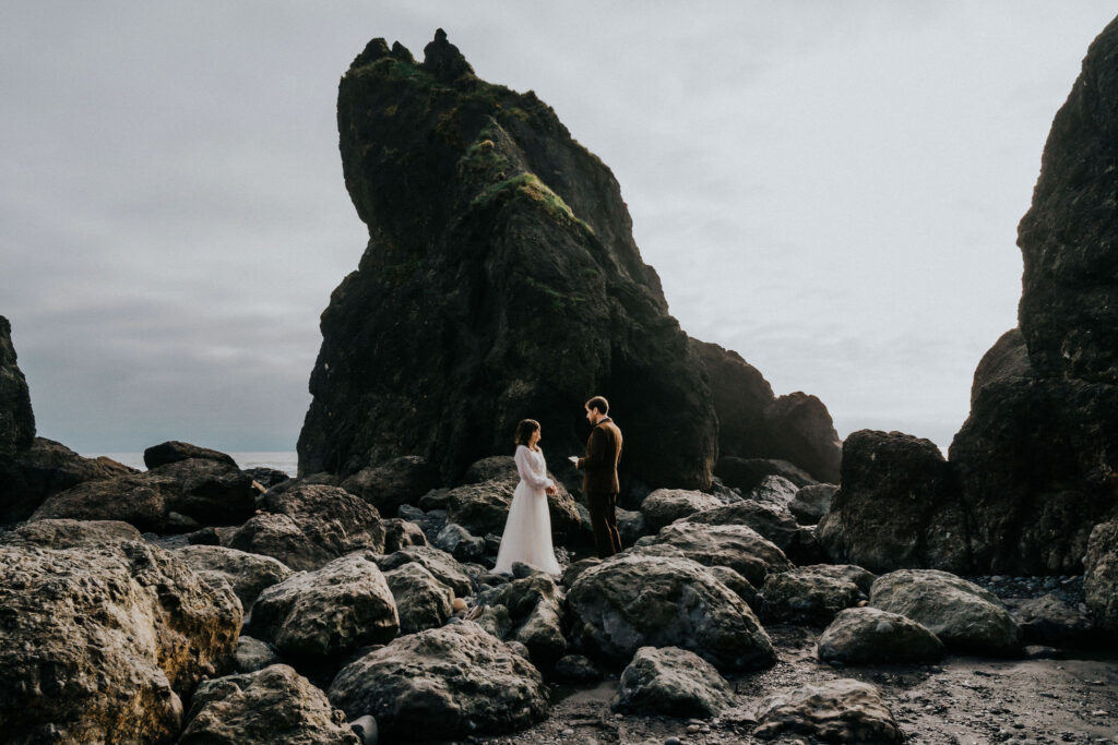 a couple in their wedding attire exchange vows on a rocky beach. the lighting is soft and the skies are moody during this spring elopement