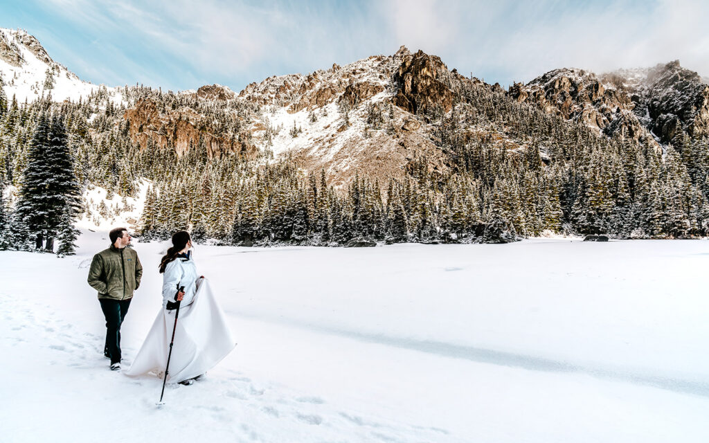 a bride and groom in their wedding attire explore a snowy landscape with trekking poles. There's fresh powder all around them and they crane their necks to see the staggering mountain views