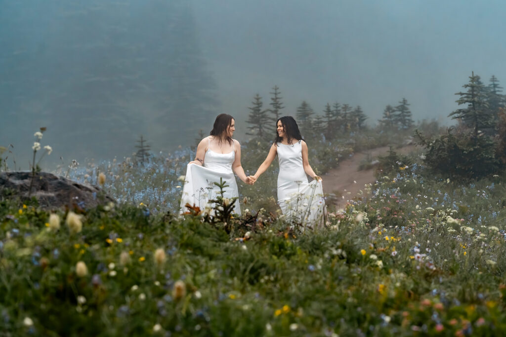 two brides walk hand in hand through a field of wildflowers smiling widely at each other in their wedding dresses